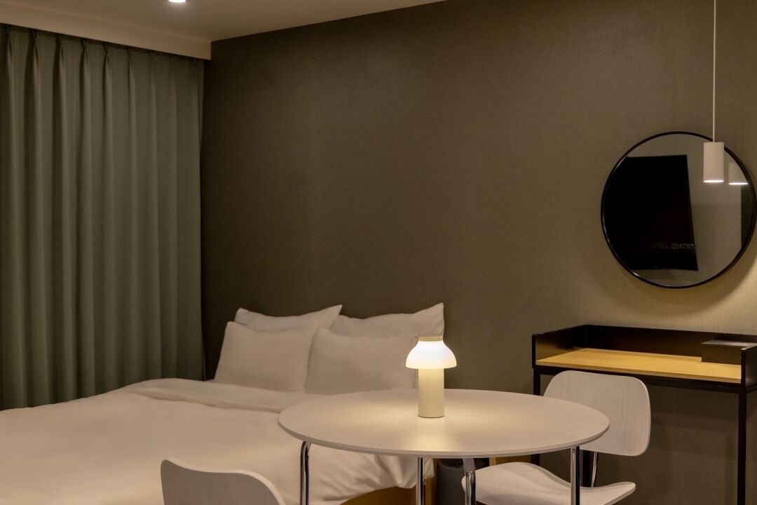 Une chambre d’hôtel avec un lit, une table ronde avec une lampe, une chaise, un bureau et un miroir au mur.