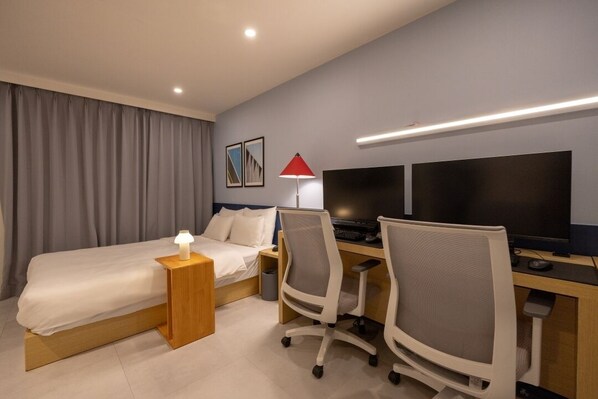A hotel room with a bed, desk, two chairs, a computer, a TV, and a lamp.