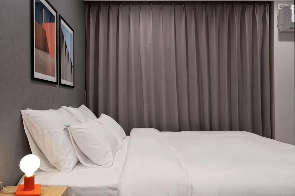 A hotel room with a bed, white bedding, a nightstand with a lamp, and a window with curtains.