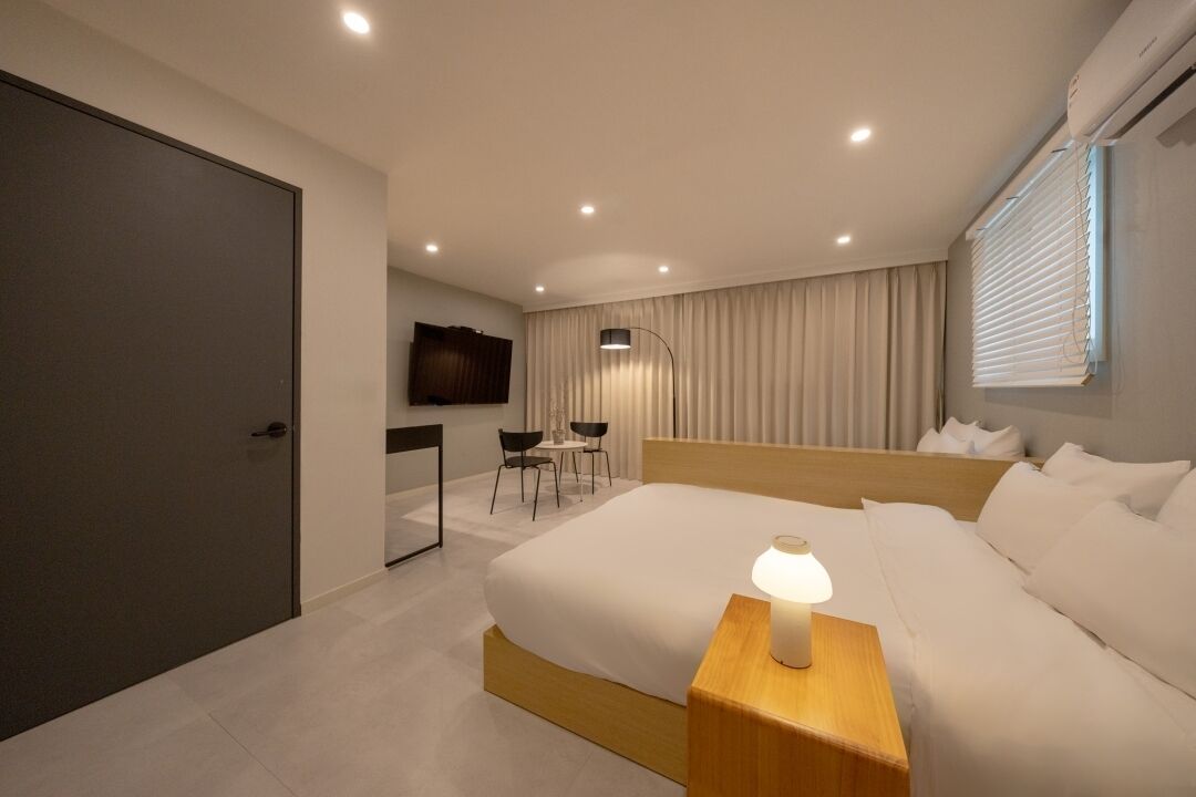 Una habitación de hotel moderna con una cama grande, una mesita de noche con lámpara, un escritorio con sillas, un televisor montado en la pared y una ventana con persianas.