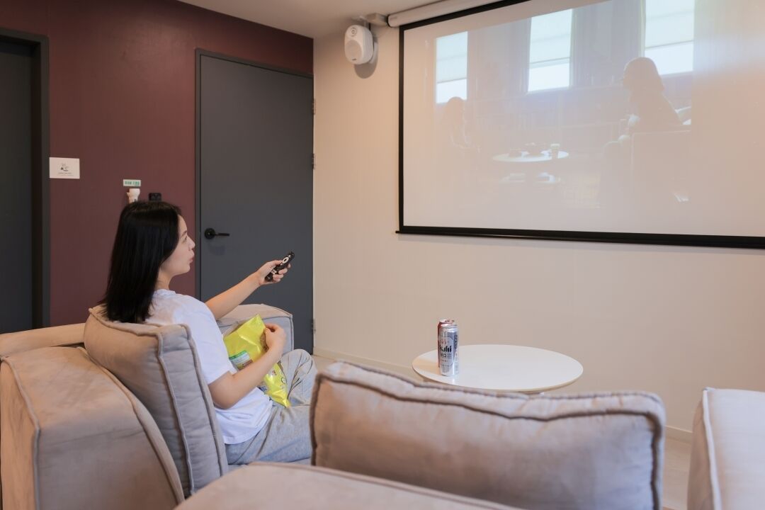 Una persona asseguda en un sofà a una habitació amb una pantalla de projector, una taula petita amb una llauna i una porta amb un pany.