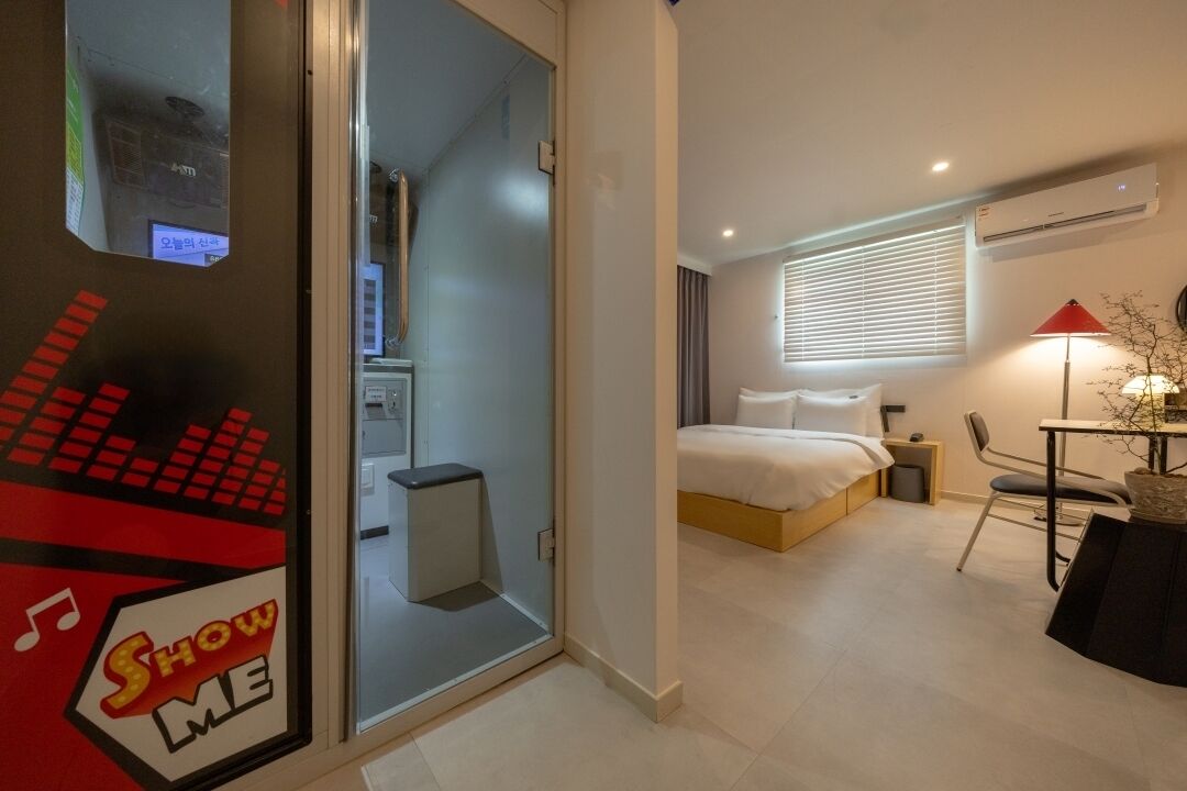 A modern hotel room with a bed, bedside table, chair, and a glass door leading to a bathroom.