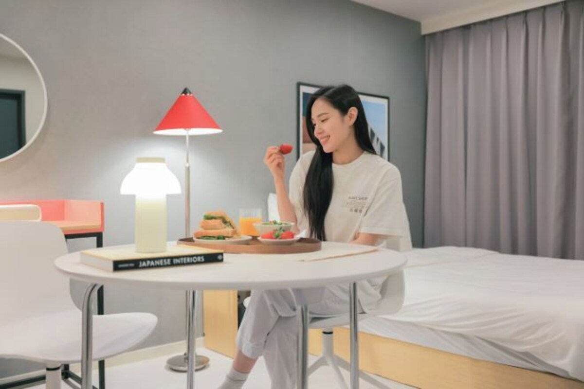A hotel room with a bed, a round table with a book titled "Japanese Interiors," a red lamp, and a person sitting at the table.