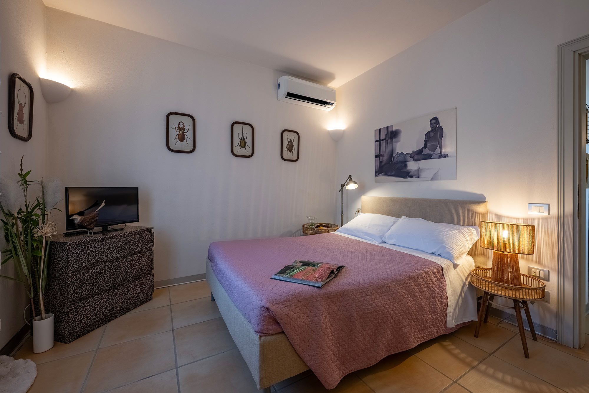 A bedroom with a bed, a television on a stand, wall-mounted air conditioning, and a painting.