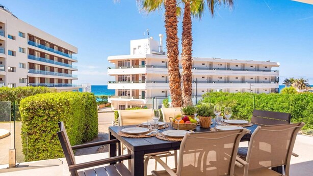 Un espace de restauration en plein air avec une table dressée pour deux personnes, entourée de chaises, de palmiers et offrant une vue sur un immeuble à plusieurs étages doté de balcons.