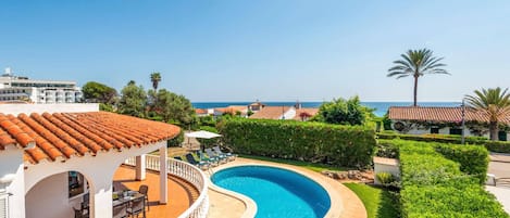 A swimming pool with a curved design, surrounded by a white fence, a covered seating area with tables and chairs, and a view of the sea.