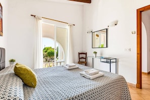 A bedroom with a bed, a chair, a small table, a mirror, and an open door leading to a balcony.