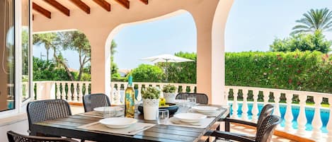 An outdoor dining area with a table set for four, surrounded by wicker chairs, overlooking a swimming pool and lush greenery.