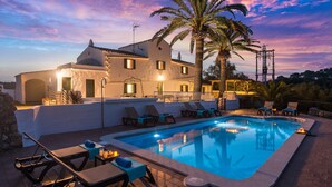 A poolside area with lounge chairs, a swimming pool, and a building with a white facade and a palm tree. - Villa Mas Alta by Villa Plus (Ferreries)