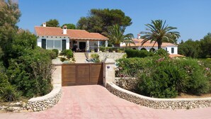 Une villa blanche avec un toit en tuiles, un chemin de pierre menant à un portail en bois, et une végétation luxuriante qui entoure la propriété.