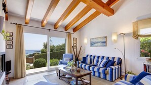 Un salon spacieux doté d’une grande fenêtre donnant sur la mer, d’un canapé à rayures bleues, d’une table basse et d’un téléviseur.