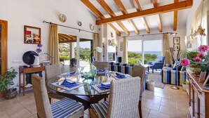 Un vaste espace de vie comprenant un coin repas avec une table pour quatre personnes, entouré de chaises en osier. De grandes fenêtres offrent une vue sur la verdure et la mer.