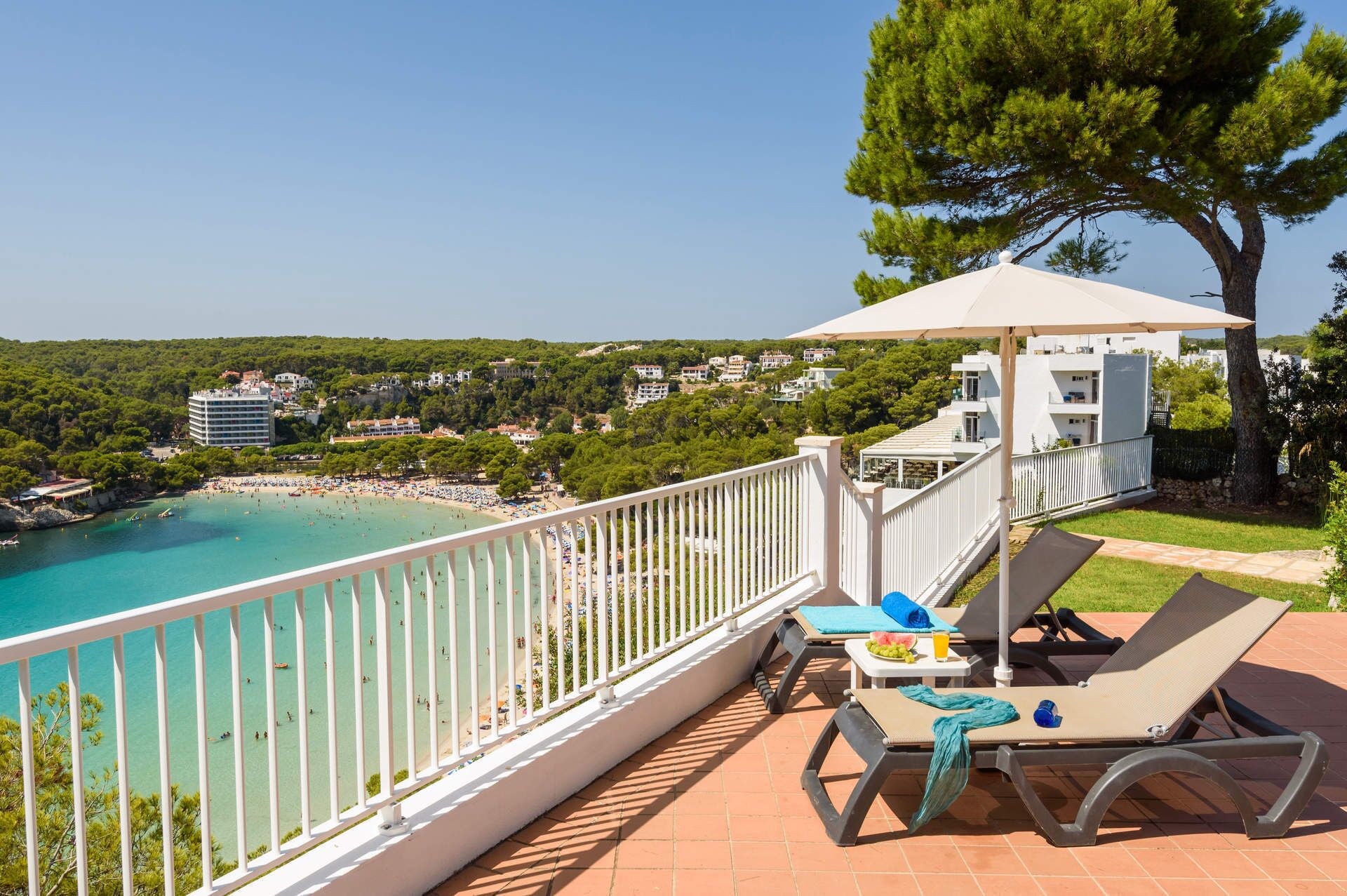 A balcony with a view of a beach and a pool, a table with a chair, and a large umbrella.