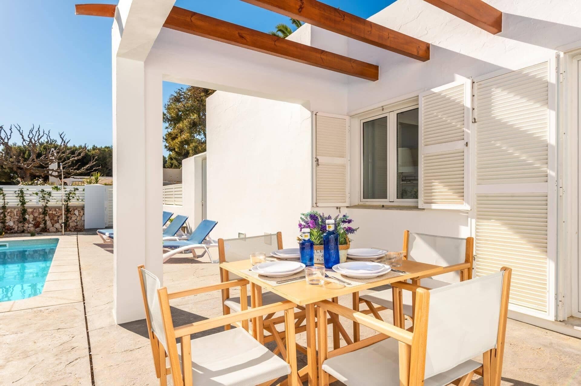 Área de refeições à beira da piscina, com móveis de madeira e uma mesa posta para dois.