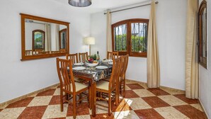 Un comedor con suelo a cuadros, sillas de madera, una mesa preparada para cuatro personas, una lámpara colgante y dos ventanas con cortinas.