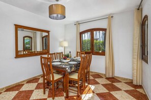 A dining area with a checkered floor, wooden chairs, a table set for four, a hanging light fixture, and two windows with curtains.