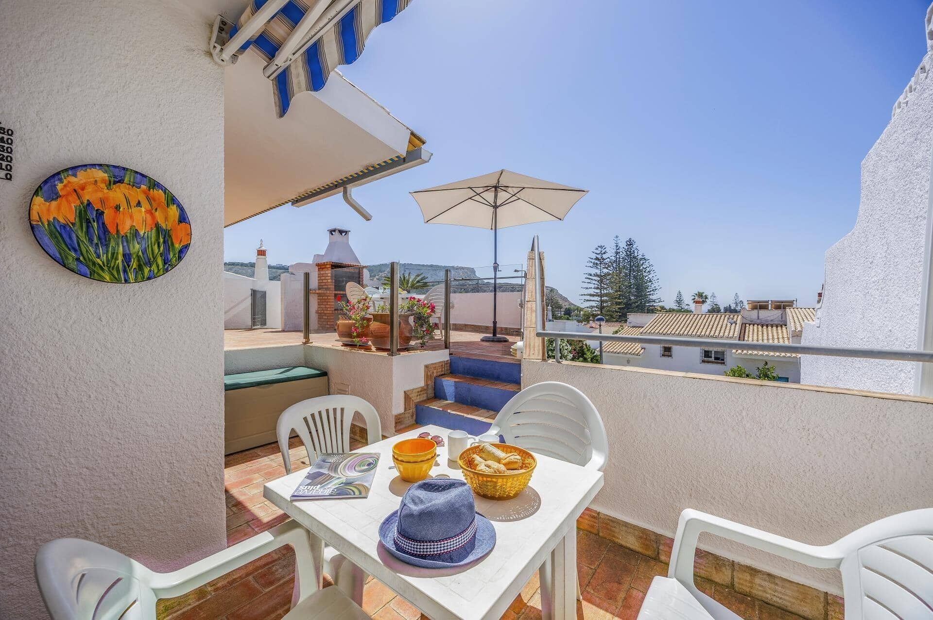 A balcony with a table set for two, a chair, a sun umbrella, and a decorative plate.