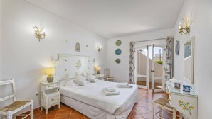 Quarto com cama, mesas de cabeceira, uma cadeira, cômoda e porta que dá para uma área externa.