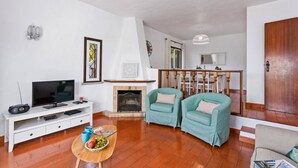 Ein Wohnzimmer mit Kamin, zwei sesseln in Türkis, einer weißen Unterhaltungsanlage und einem Essbereich mit Holztisch und Stühlen.