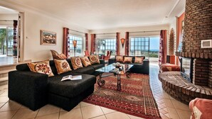 A spacious living room with a large black sectional sofa, a glass coffee table, and a red patterned rug. There is a brick fireplace and a large window with curtains. - Villa Nenufa by Villa Plus (Albufeira)