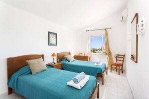 A hotel room with two beds, a window with a view, a wooden chair, and a small table with a lamp.