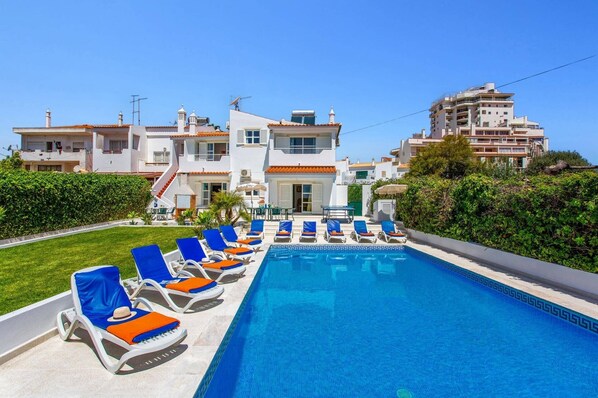 A swimming pool with lounge chairs, a building with balconies, and a clear sky.