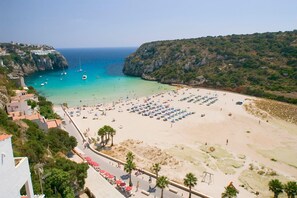 A beach with rows of sunbeds and umbrellas, a clear blue sea, and rocky cliffs in the background. - Villa Limon by Villa Plus (Cala en Porter)