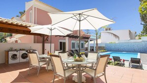 Eine Terrasse mit einem runden Tisch, Stühlen und einem großen Sonnenschirm. Im Hintergrund ist ein Pool und ein Gebäude mit einer rosafarbenen Fassade zu sehen.