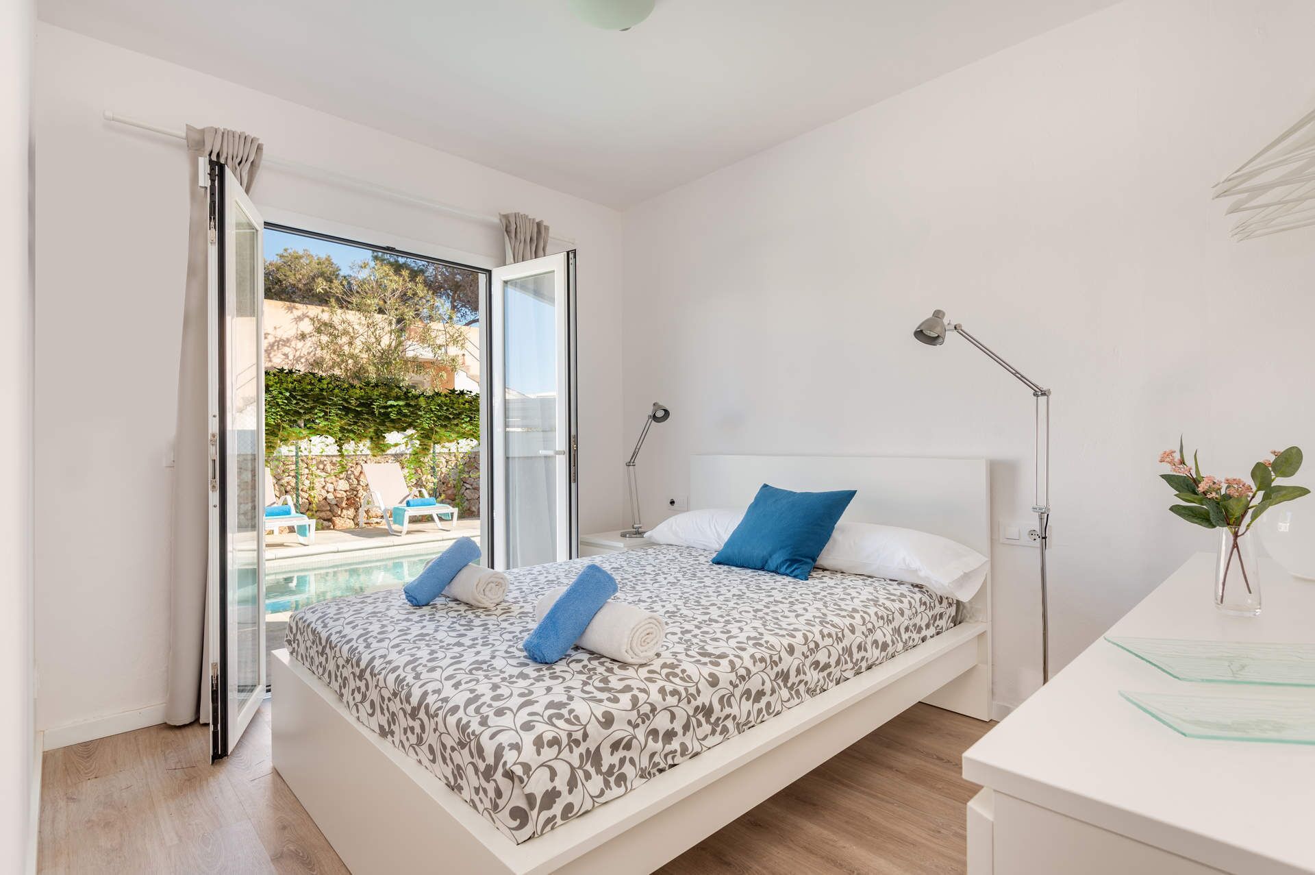 A bedroom with a bed, bedside lamps, a desk, and a view of a pool and outdoor seating.