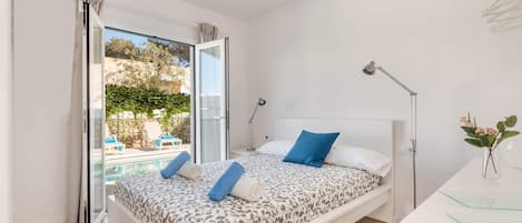A bedroom with a bed, bedside lamps, a desk, and a view of a pool and outdoor seating.