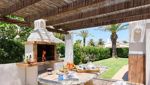 An outdoor patio with a stone table, a stone bench, a brick oven, and a pergola with a woven roof. - Villa Carianne by Villa Plus (Son Carrió)