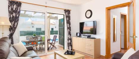 Een moderne woonkamer met een bank, een salontafel, een televisie en een glazen schuifdeur die leidt naar een balkon met buitenmeubels.
