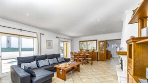 A spacious living area with a sofa, dining table, and chairs, a kitchen area with wooden cabinets, and large windows offering a view. - Villa Anamar by Villa Plus (Albufeira)