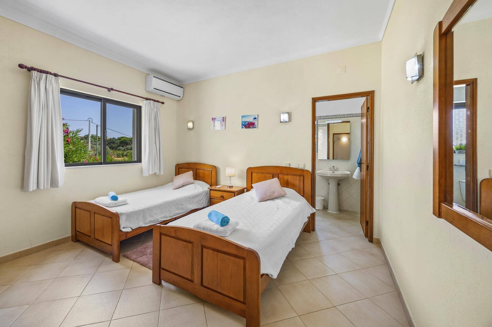 A hotel room with two wooden beds, a window with curtains, an air conditioning unit, a bedside table with a lamp, a small wall-mounted shelf, and a bathroom with a sink and mirror.