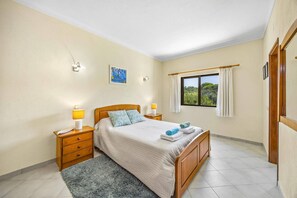 A bedroom with a bed, two bedside lamps, a nightstand, a window with curtains, and a painting on the wall. - Villa Horta by Villa Plus (Lagoa)