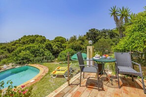 A poolside area with a table and chairs, surrounded by lush greenery and a clear blue sky.