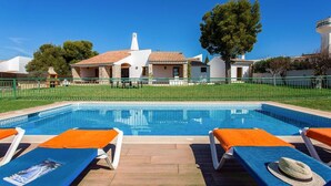 Une piscine entourée de chaises longues, un bâtiment blanc avec un toit en tuiles et un grand arbre en arrière-plan.