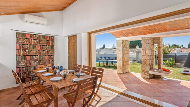 Un coin repas avec une table en bois dressée pour six personnes, une bibliothèque et une vue sur une piscine et un jardin.