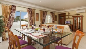 A dining area with a glass table, purple chairs, and a sofa. There are curtains, a television, and a cabinet with books. - Villa Cubos by Villa Plus (Albufeira)