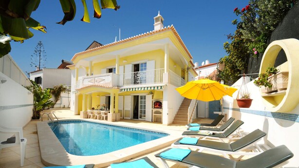 A pool area with lounge chairs and a yellow umbrella, a two-story building with balconies, and a clear blue sky. - Villa Cubos by Villa Plus (Albufeira)