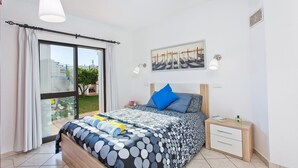 Ein Schlafzimmer mit großem Fenster, einem Bett mit gemustertem Überwurf, einem Nachttisch und einem an der Wand hängenden Bild.