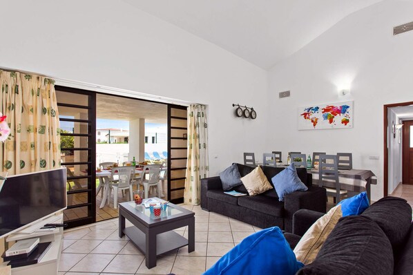 A living room with a sofa, coffee table, TV, and a dining area with chairs and a table. - Villa Neptuno Sol by Villa Plus (Albufeira)