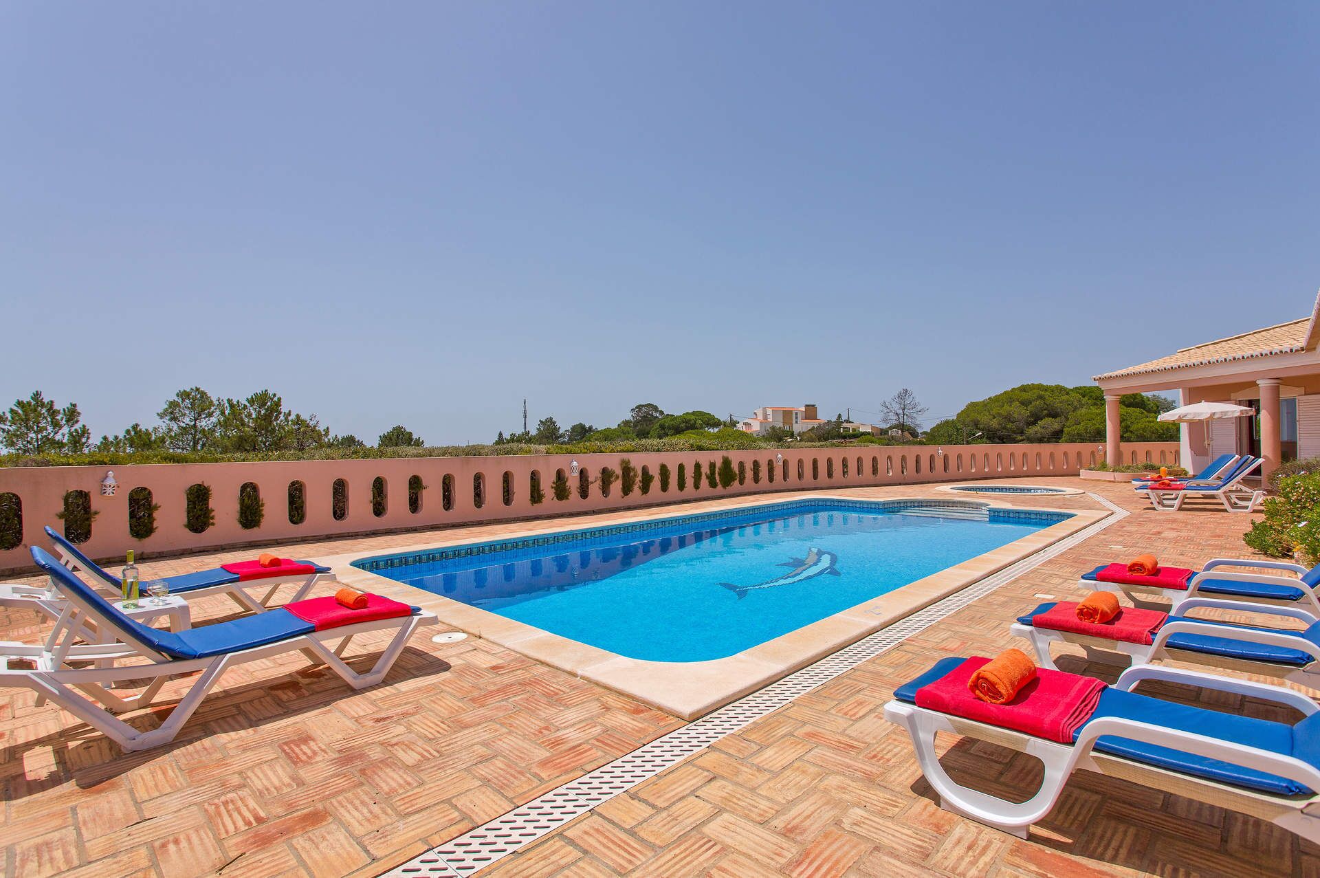 Una piscina con acqua azzurra, circondata da sdraio con cuscini rossi e blu, e un'area patio piastrellata.