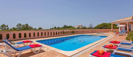 Una piscina con acqua azzurra, circondata da sdraio con cuscini rossi e blu, e un'area patio piastrellata.