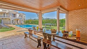 A covered outdoor dining area with a pool and a view of a large house in the background. - Villa Tess by Villa Plus (Praia do Carvoeiro)