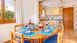 A dining area with a wooden table set for four, blue placemats, and chairs. A kitchen with wooden cabinets and a tiled backsplash is visible in the background. - Villa Clara by Villa Plus (Binibeca)