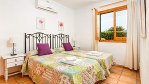 A bedroom with two beds, a window with curtains, a bedside table with a lamp, and a wall-mounted air conditioner. - Villa Clara by Villa Plus (Binibeca)