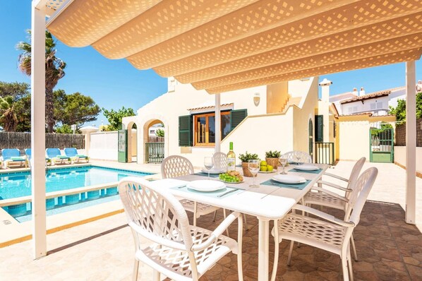 Un espace repas aménagé au bord de la piscine, avec une table dressée pour deux personnes, une bouteille de vin et un panier de fruits.