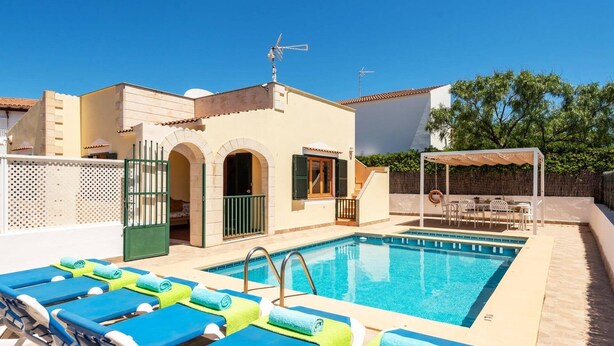 Un área de piscina con sillones reclinables, una zona de estar techada y un edificio con un arco abierto.