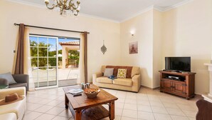 A living room with a sofa, a coffee table, a TV stand, and a balcony with a view of greenery. - Villa Arvela by Villa Plus (Vale Rebelho)
